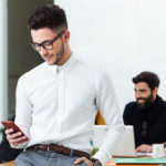 A man checks his cell phone while colleagues work on laptops.