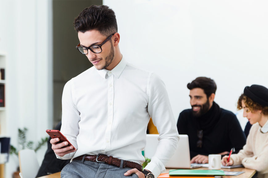 A man checks his cell phone while colleagues work on laptops.